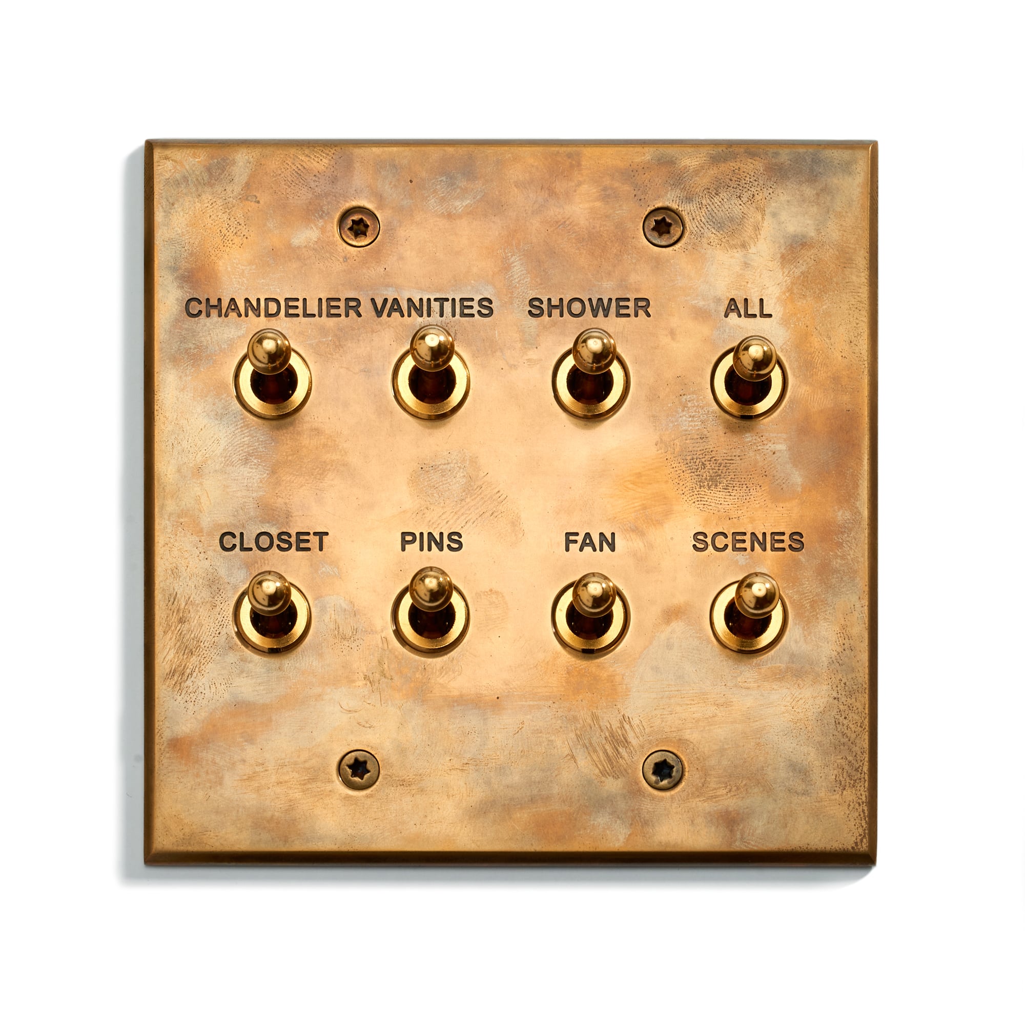 A brass light switch panel with eight toggle switches labeled: "Chandelier Vanities," "Shower," "All," "Closet," "Pins," "Fan," and "Scenes." The surface appears aged and tarnished.