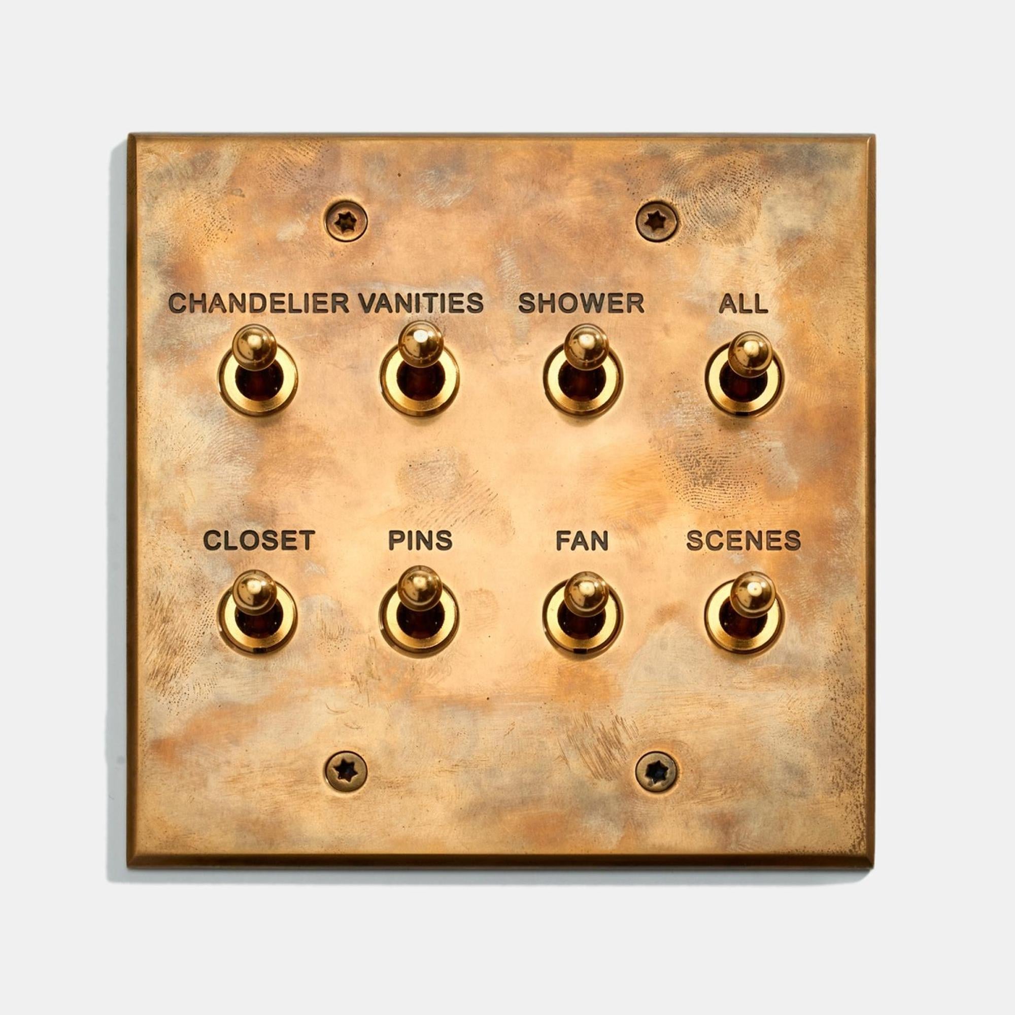 A brass switch panel with eight toggle switches labeled: "Chandelier," "Vanities," "Shower," "All," "Closet," "Pins," "Fan," and "Scenes." The panel has a worn, vintage appearance.