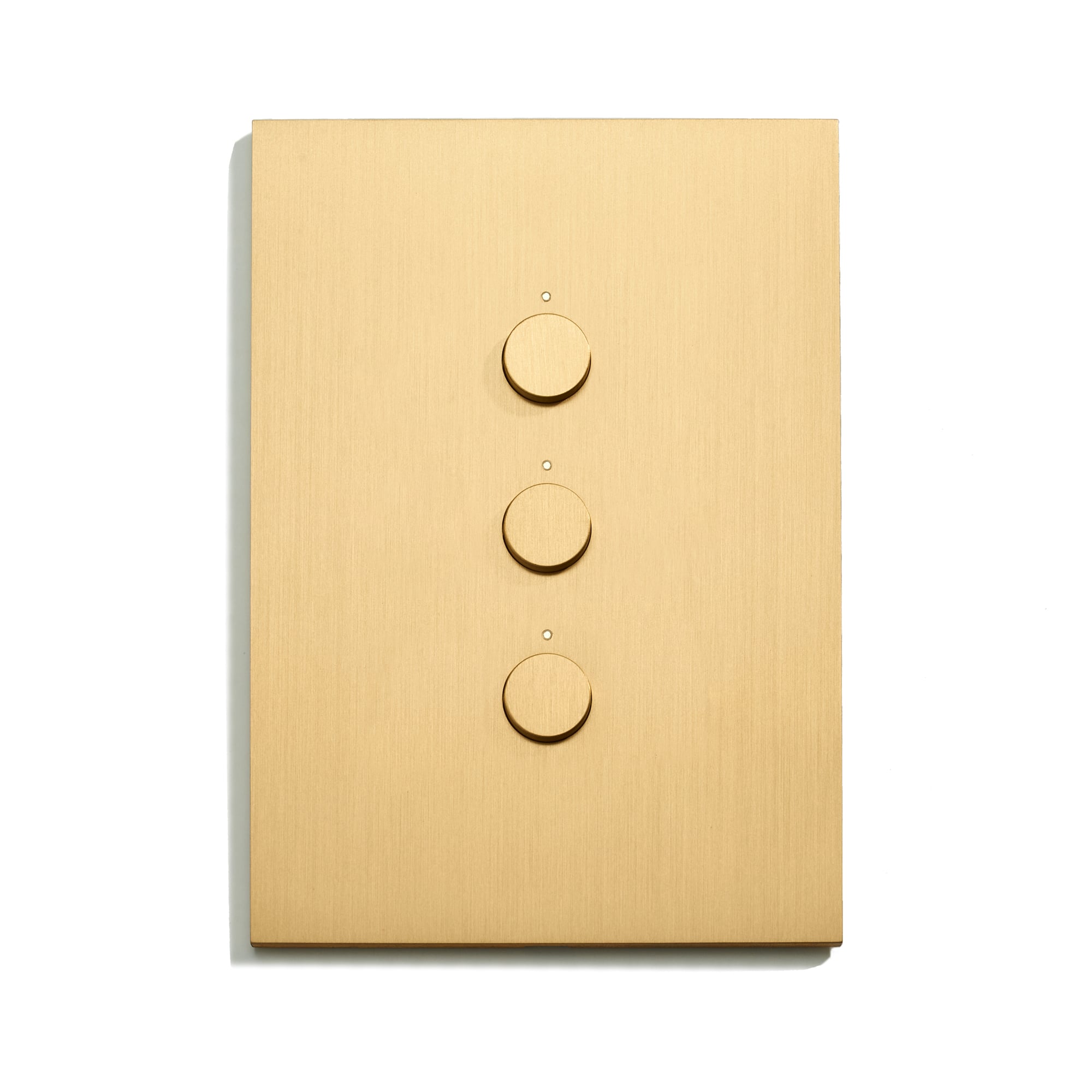 A rectangular, brass-colored light switch panel with three circular dials arranged vertically in the center. Each dial has a small dot above it. The panel has a sleek, minimalist design and casts a slight shadow to the left on a white background.