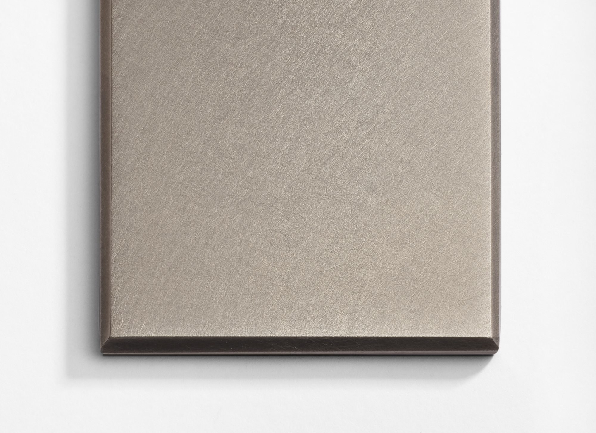 Close-up of the corner of a brushed metal sheet with a smooth, matte finish, placed on a white background. The surface has a subtle linear texture, and the edges are sharply defined.