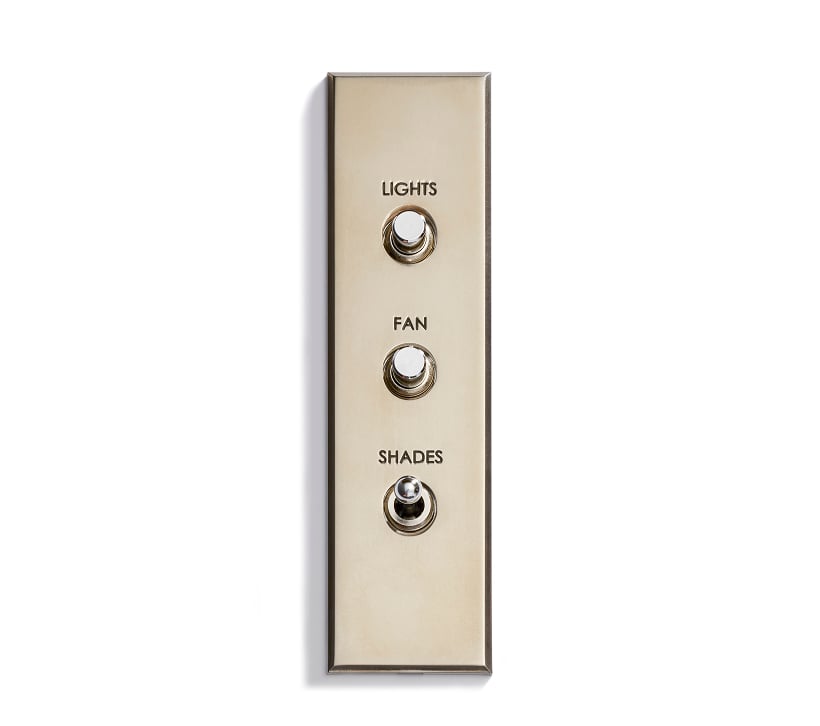 A metallic switch panel with three toggle switches labeled from top to bottom: "LIGHTS," "FAN," and "SHADES," mounted on a white wall.