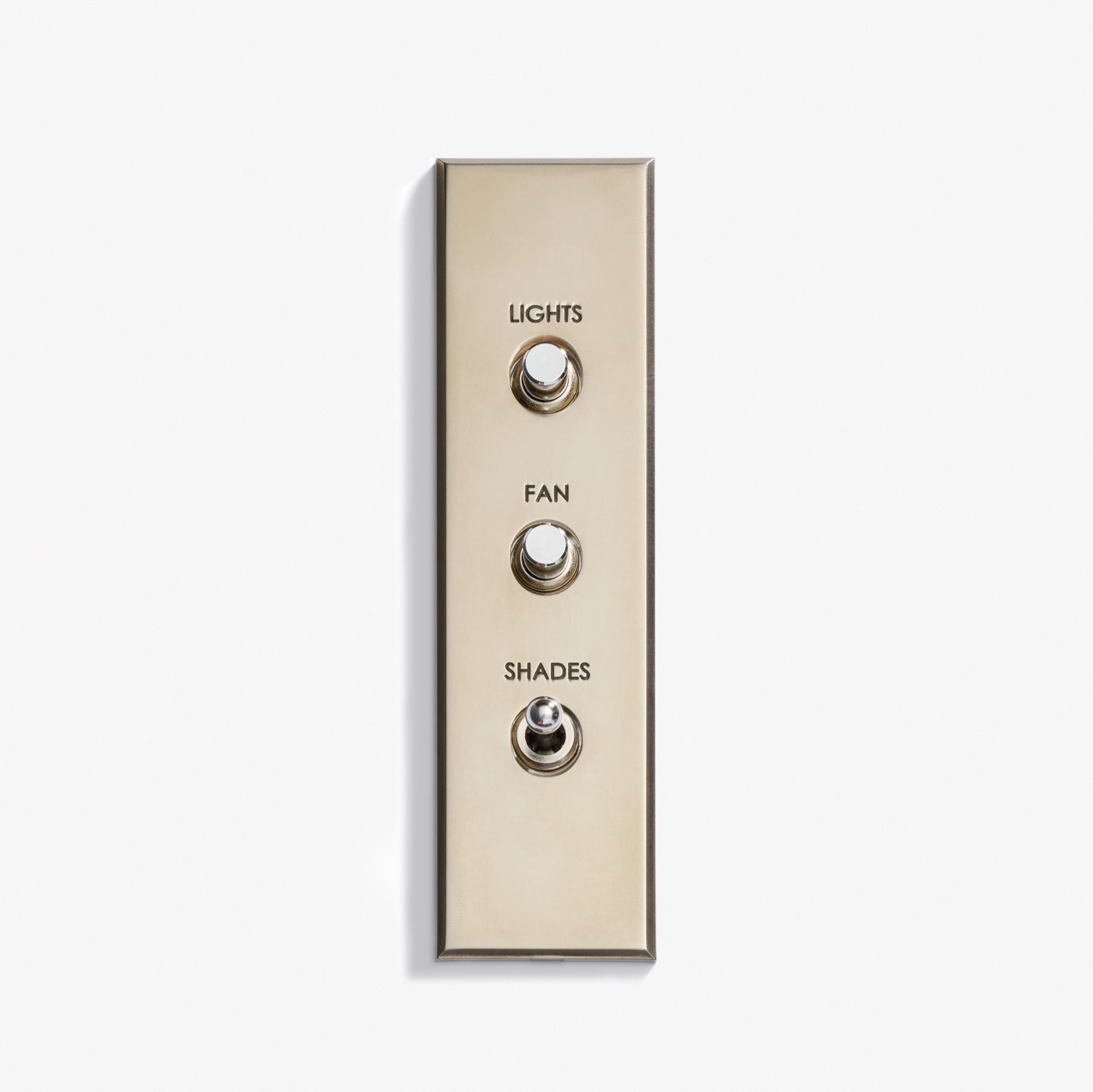 A Luxury Classique Keypad 38 x 133 brass switch panel with three toggle switches labeled "LIGHTS," "FAN," and "SHADES" sits on a white background. All switches are down except the bottom one, which is up.