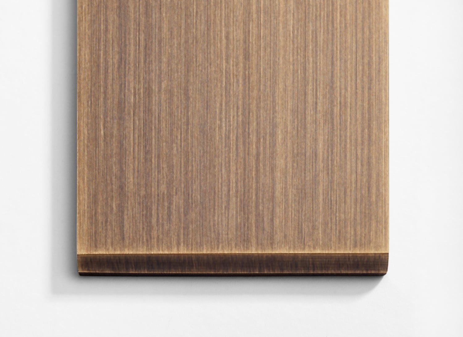 A close-up of a brown wooden board with a distinct grain pattern. The edges of the board are smooth, and the bottom edge has a rounded finish. The board is placed on a white surface, and only a portion of the board is visible in the image.