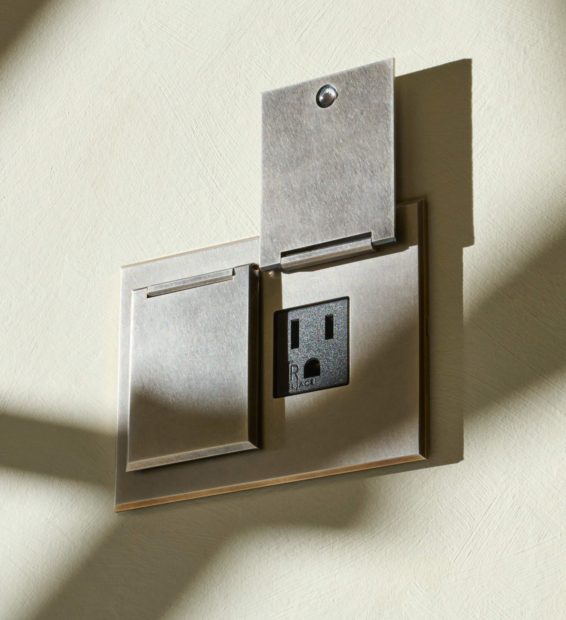 A metallic wall plate with a closed switch cover on the left and an uncovered black electrical outlet on the right, both casting shadows on a textured light-colored wall.