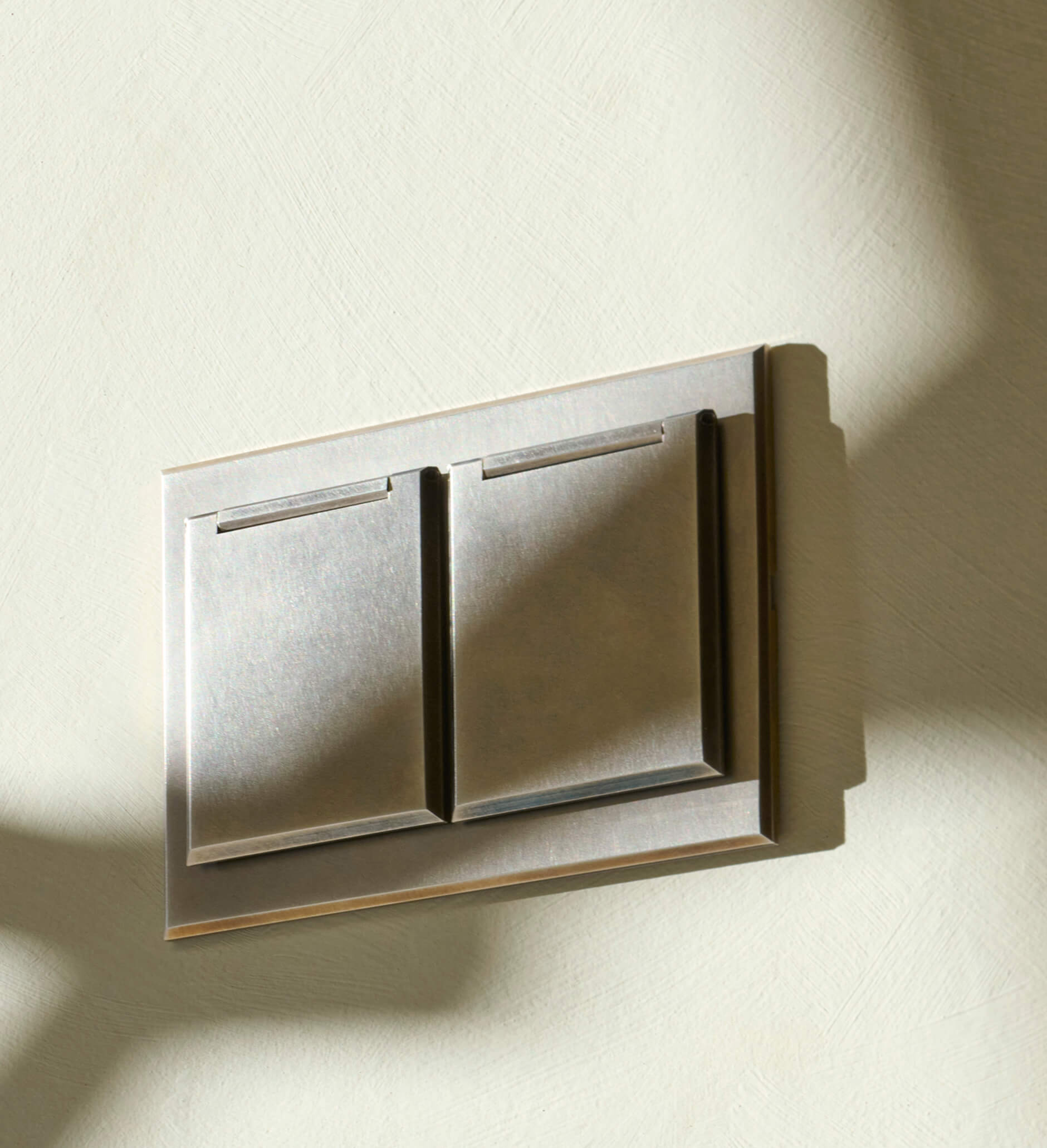 A close-up of a modern, metallic double light switch mounted on a textured, light-colored wall. Shadows fall diagonally across the scene.