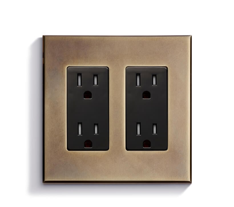 A brass-finished wall plate with two black duplex electrical outlets is mounted on a white wall, photographed from the front.