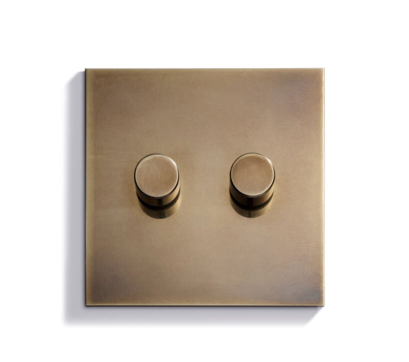 A square brass light switch plate with two round, matching brass dimmer knobs set against a white background.