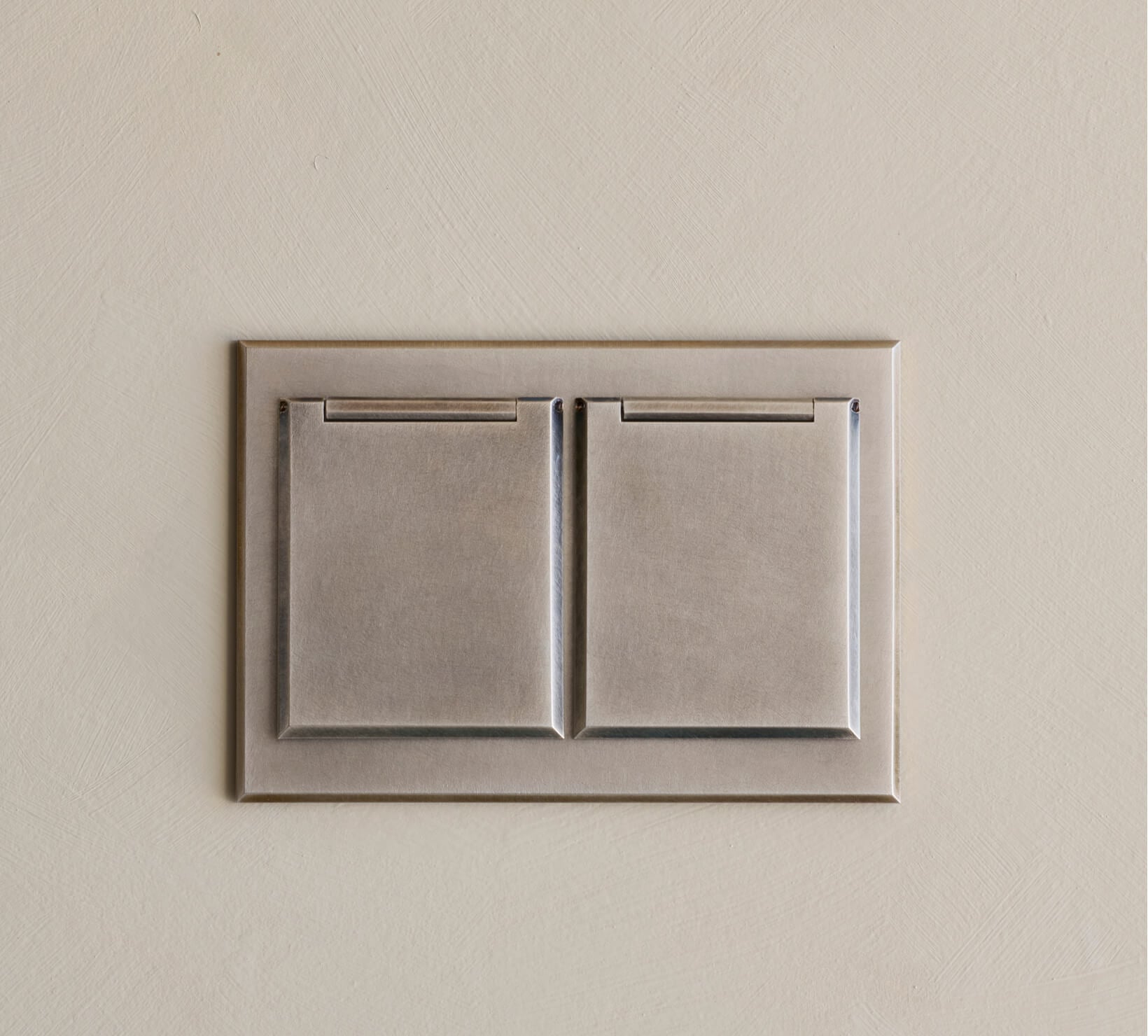 A close-up of a metallic double light switch with a modern, rectangular design mounted on a beige wall.
