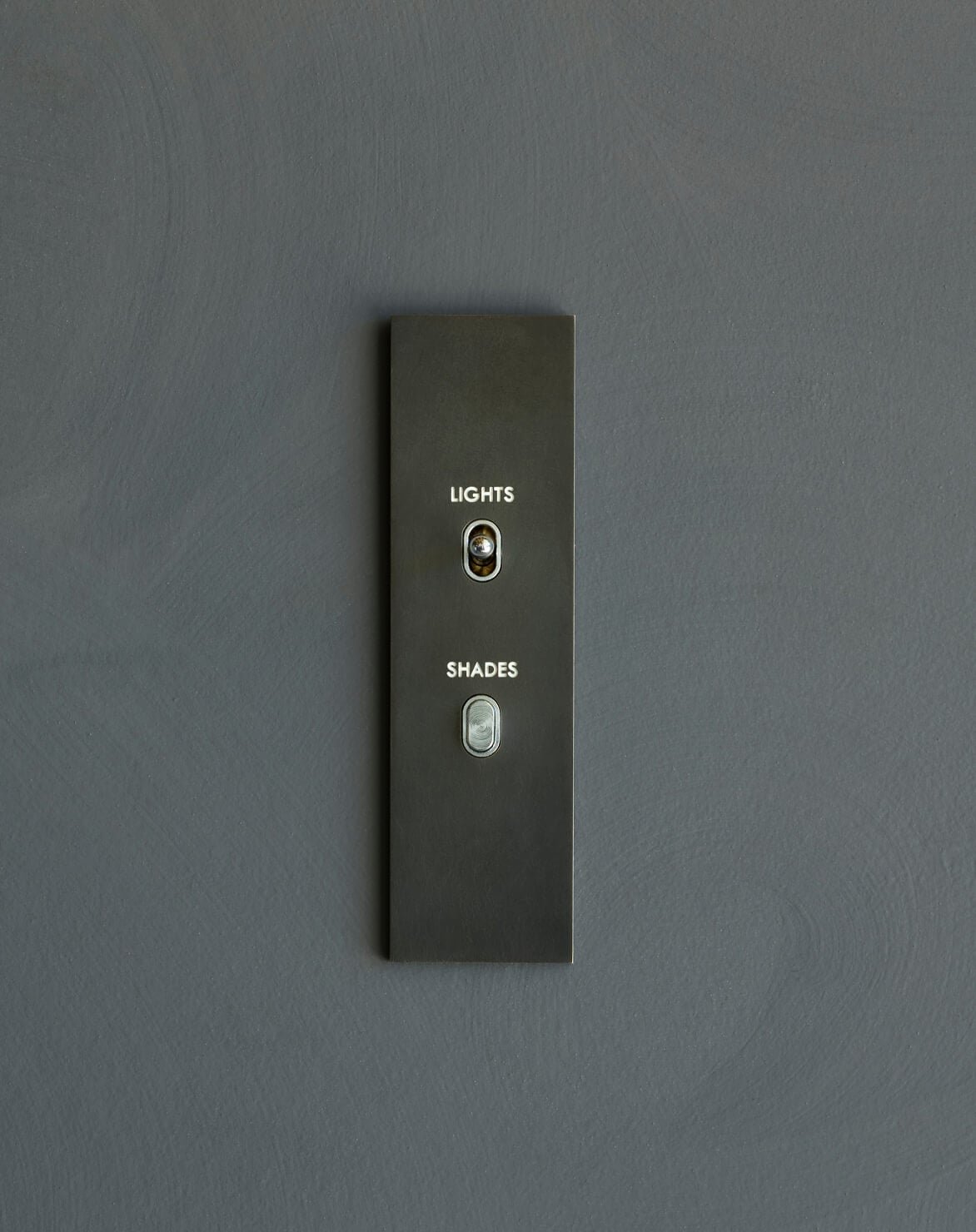 A minimalist wall panel with two buttons labeled "LIGHTS" and "SHADES" is mounted on a plain gray wall.