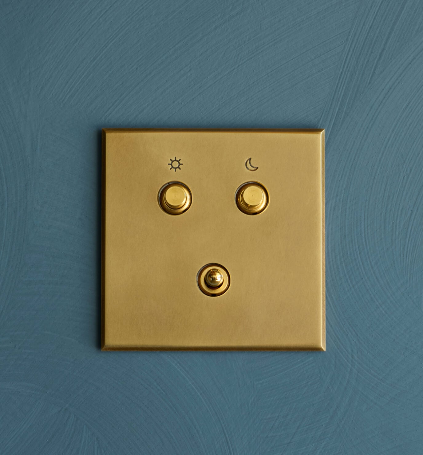 A gold light switch panel with three round switches on a textured teal wall; the top left switch has a sun symbol, the top right has a moon symbol, and the bottom switch is unmarked.
