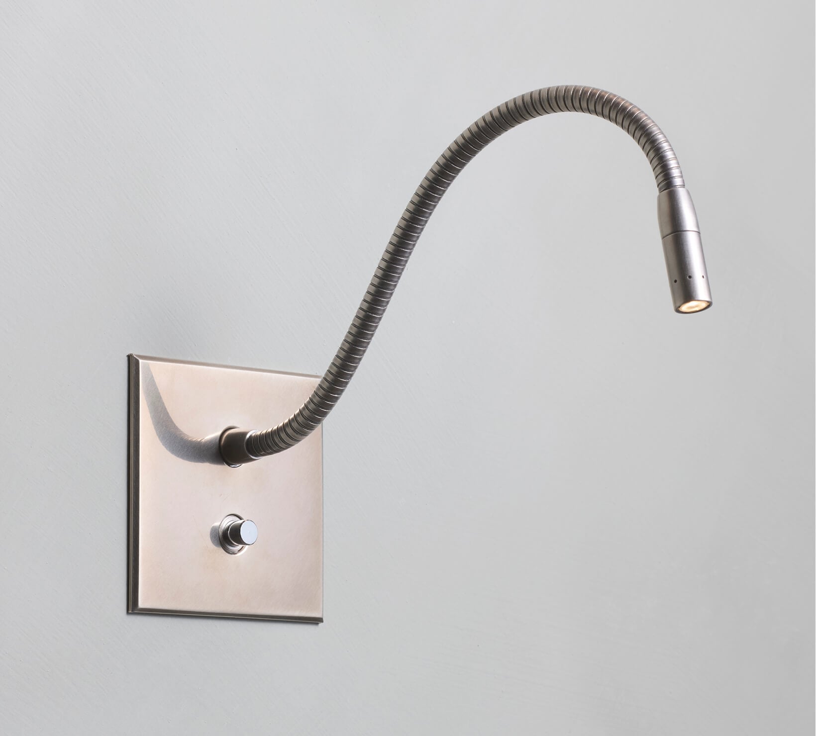 A wall-mounted, flexible-neck reading light with a metallic finish is attached to a rectangular base plate with a round on/off button below the light on a plain light gray wall.