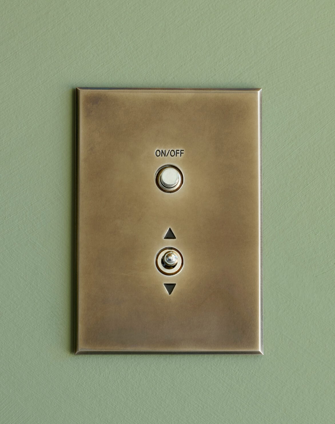 A brass light switch plate with two round buttons labeled "ON/OFF" and an arrow pointing up and down, mounted on a green wall.